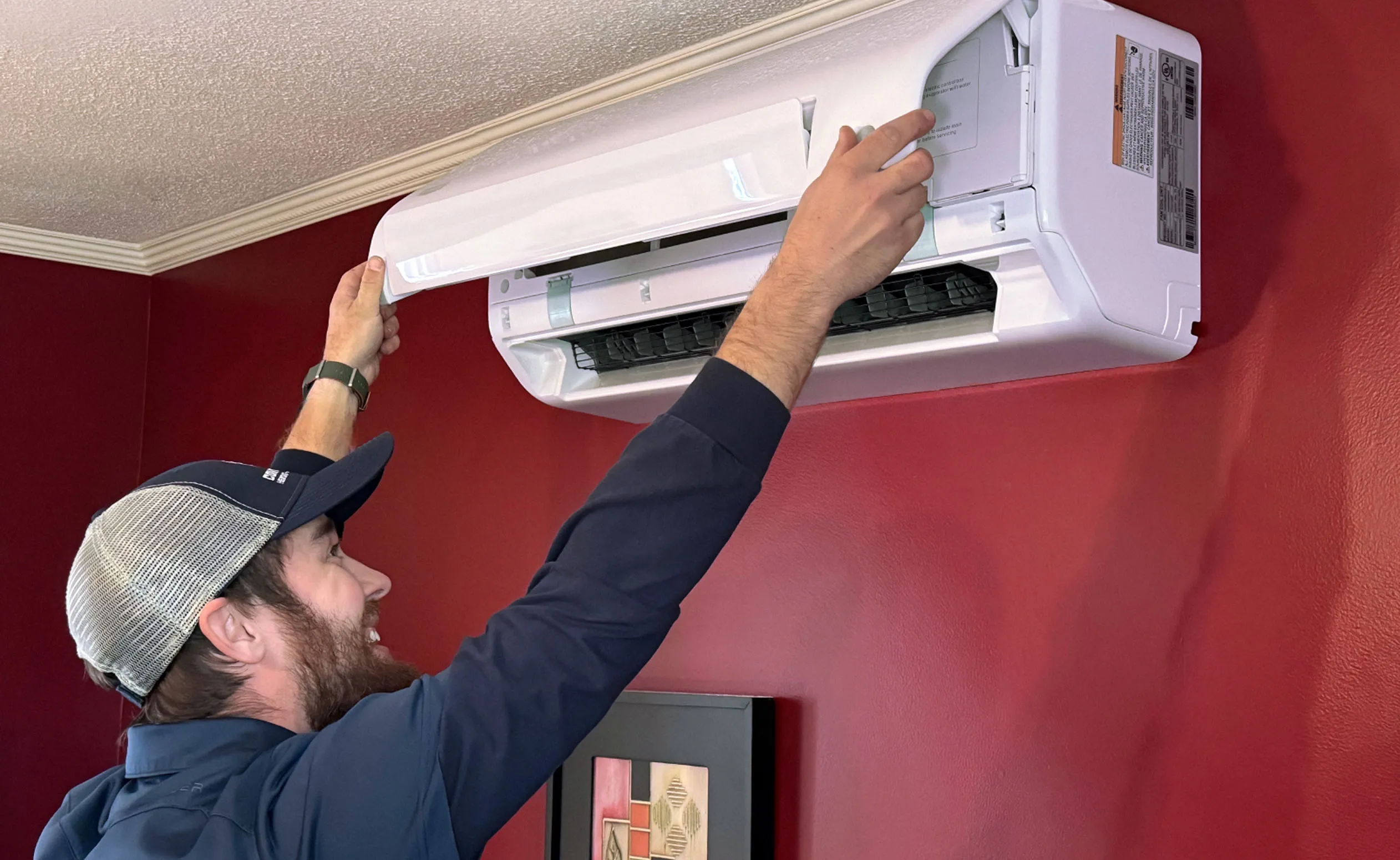 Technician inspecting and servicing a Mitsubishi Electric outdoor air conditioning unit next to a brick wall.